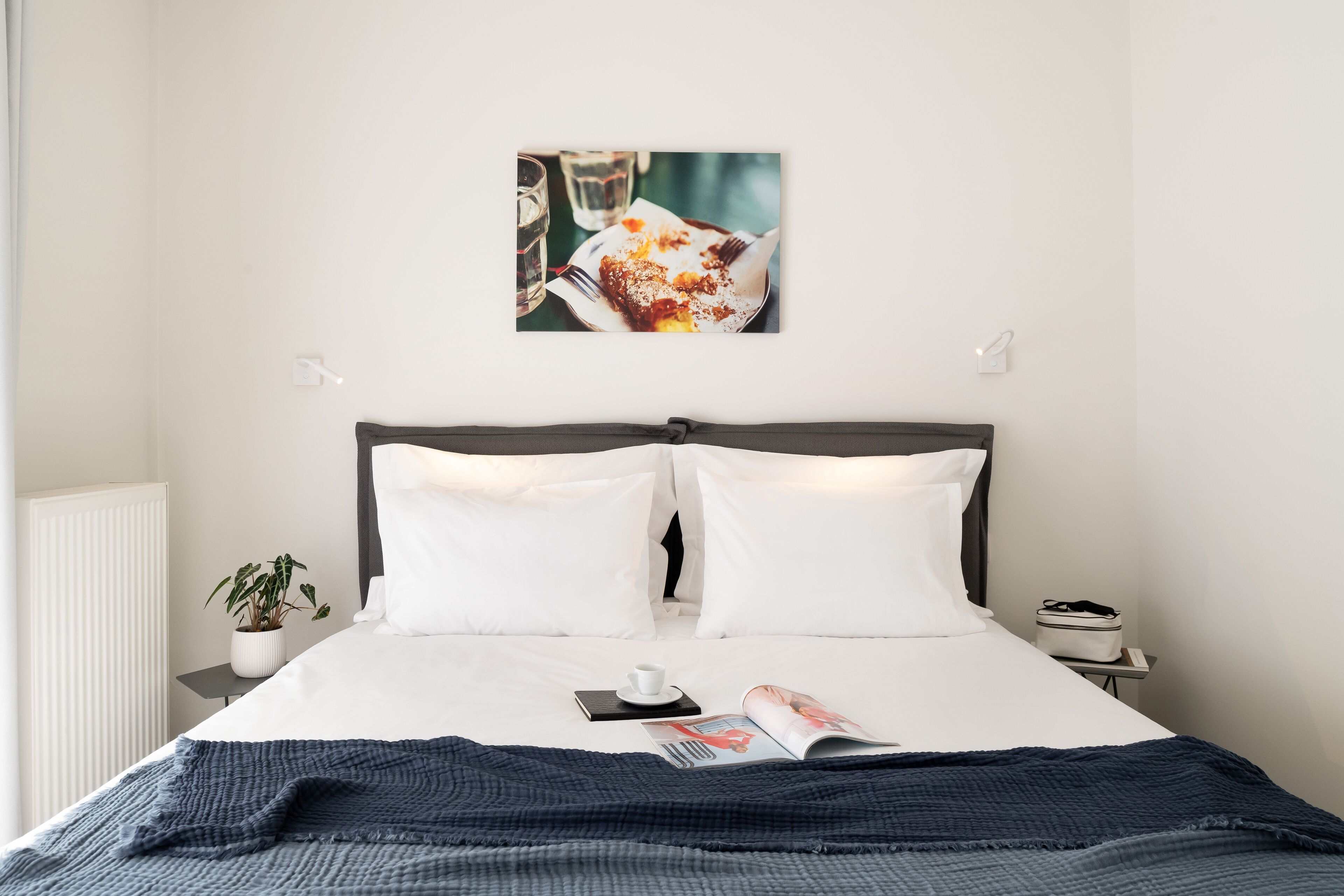 Un letto rifatto con lenzuola bianche, una coperta blu scuro, una rivista, una tazza e una piccola pianta in vaso su un comodino.