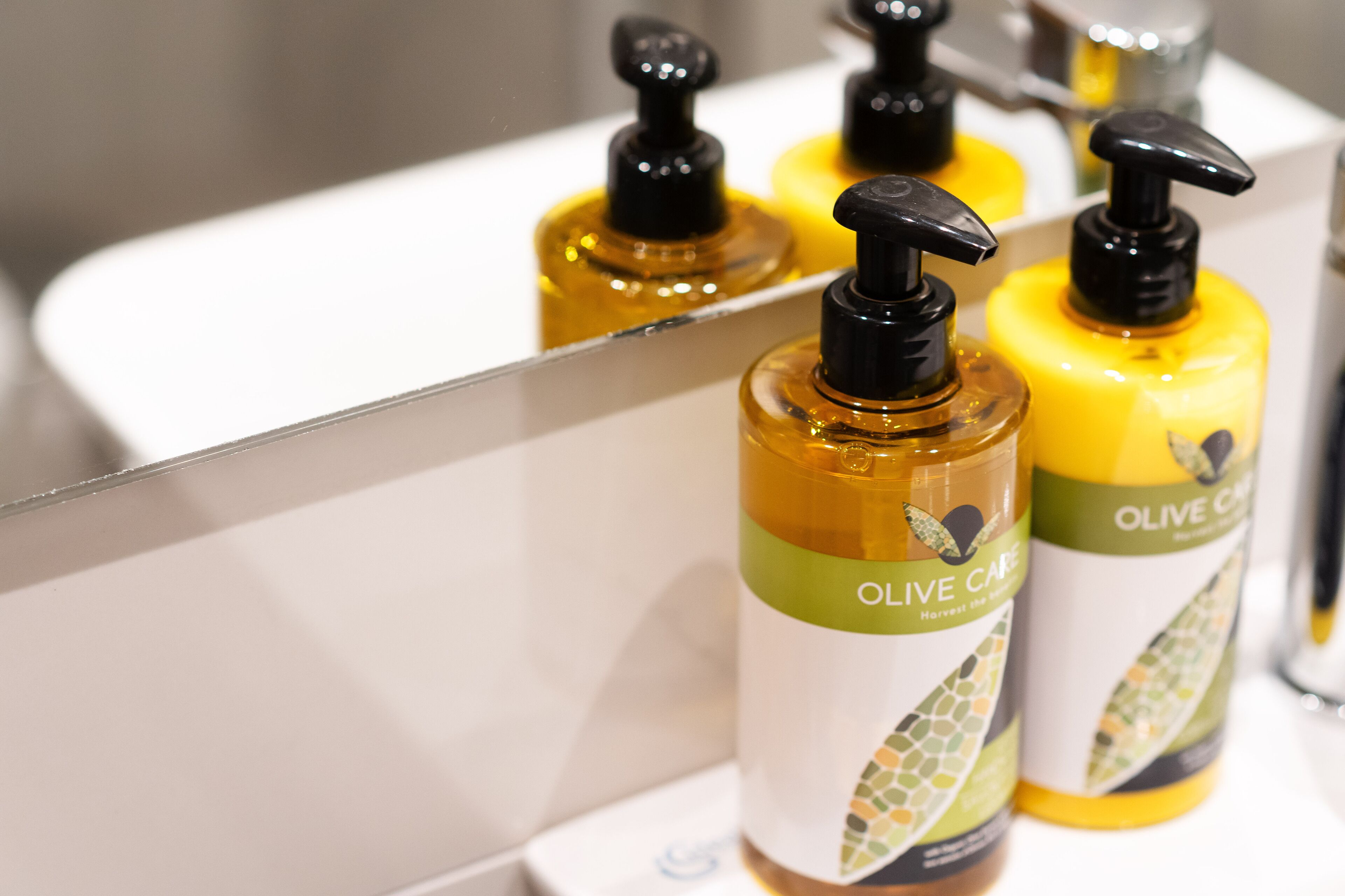 A bathroom with a white sink and mirror. There are three bottles of olive oil soap on the sink.
