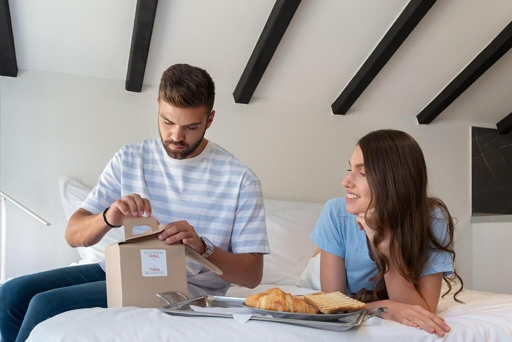 A man and a woman sitting on a bed with a box of bread and a tray of croissants.