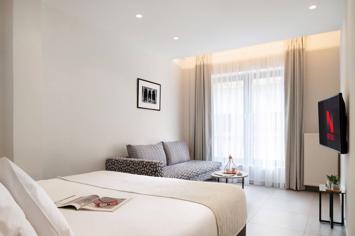 A modern hotel room with a large bed, a small sofa, a TV, and a window with curtains.
