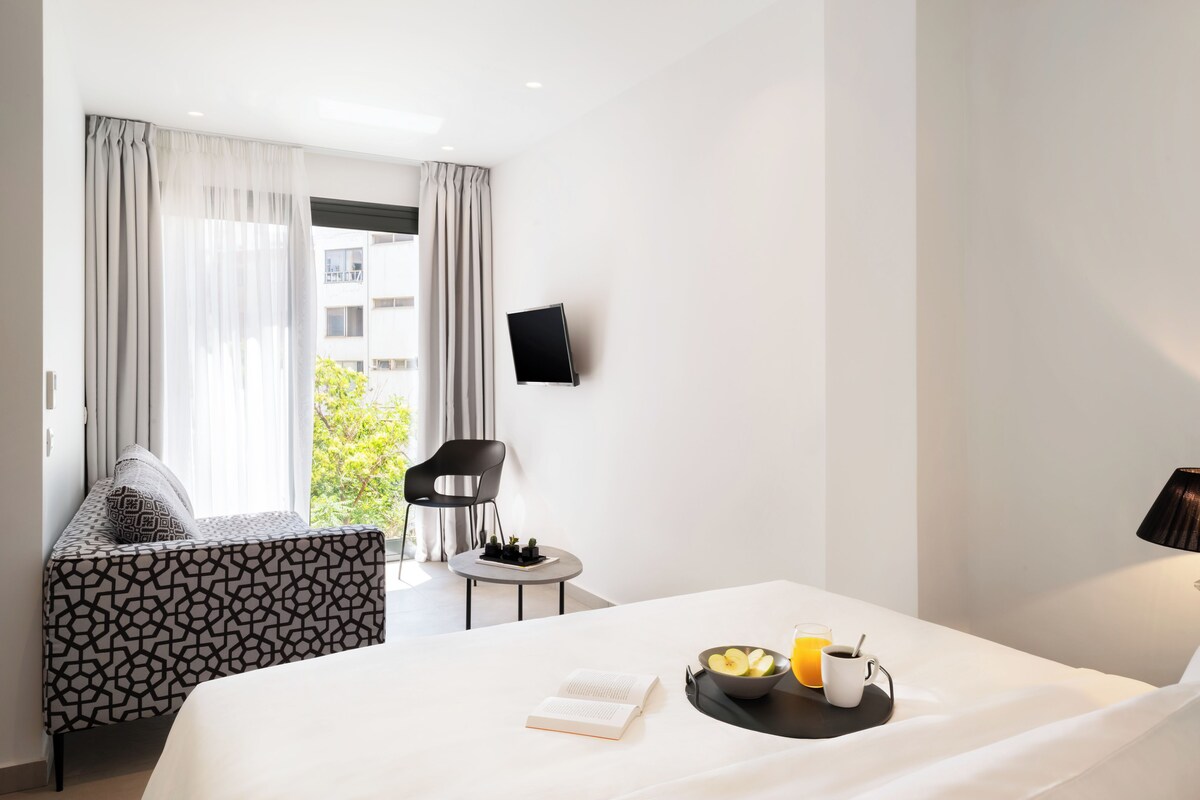 A hotel room with a bed, a small table with a bowl of fruit and a cup, a chair, a TV on the wall, and a window with curtains.