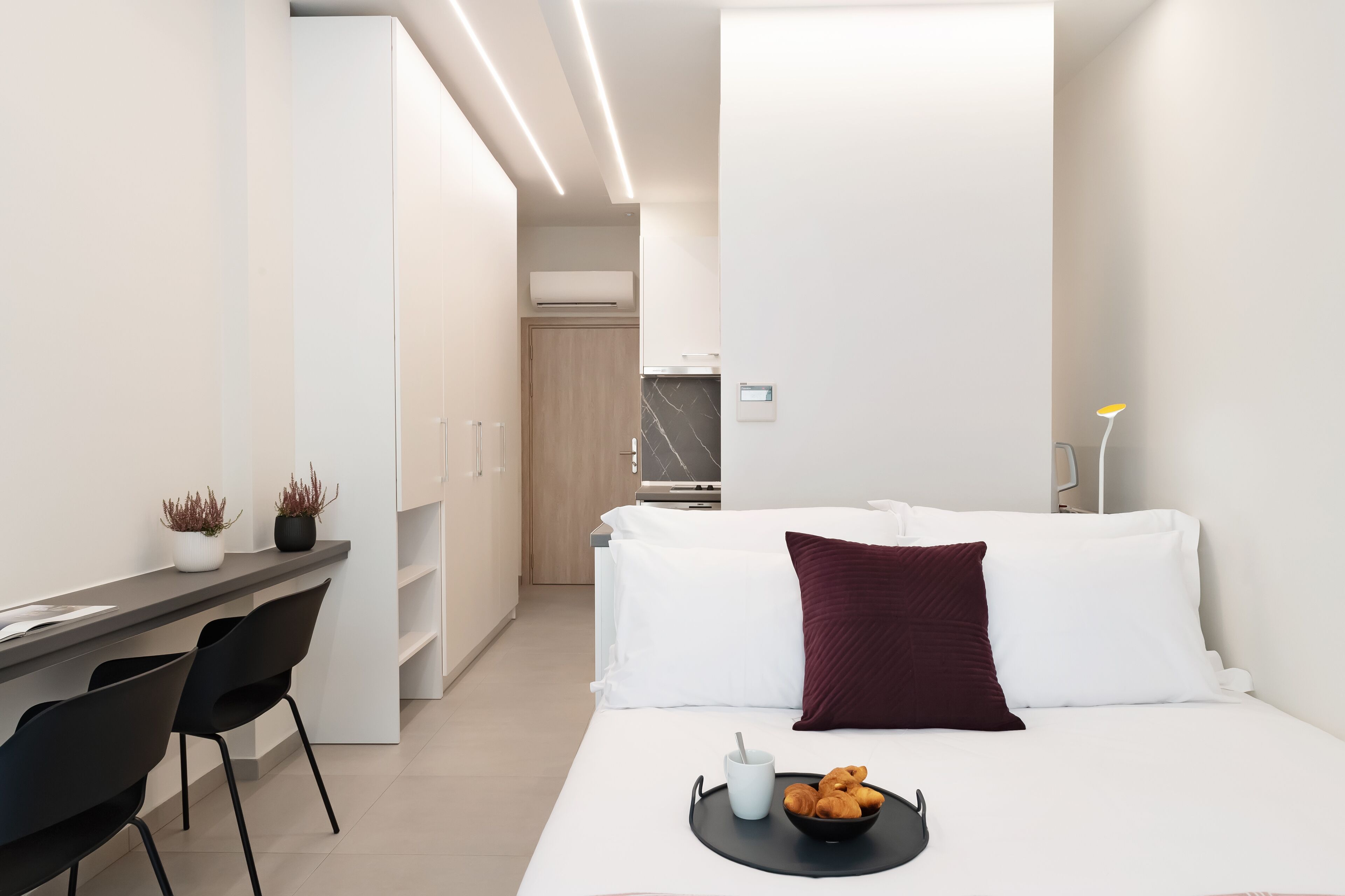 A modern hotel room with a bed, a desk with chairs, a small kitchen area, and a bedside table with a tray of croissants.