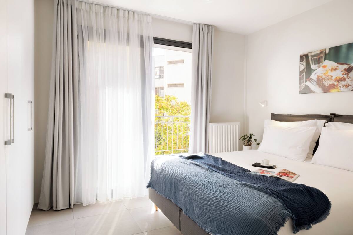 A modern hotel room with a large bed, a window with sheer curtains, a view of greenery outside, and a painting on the wall.