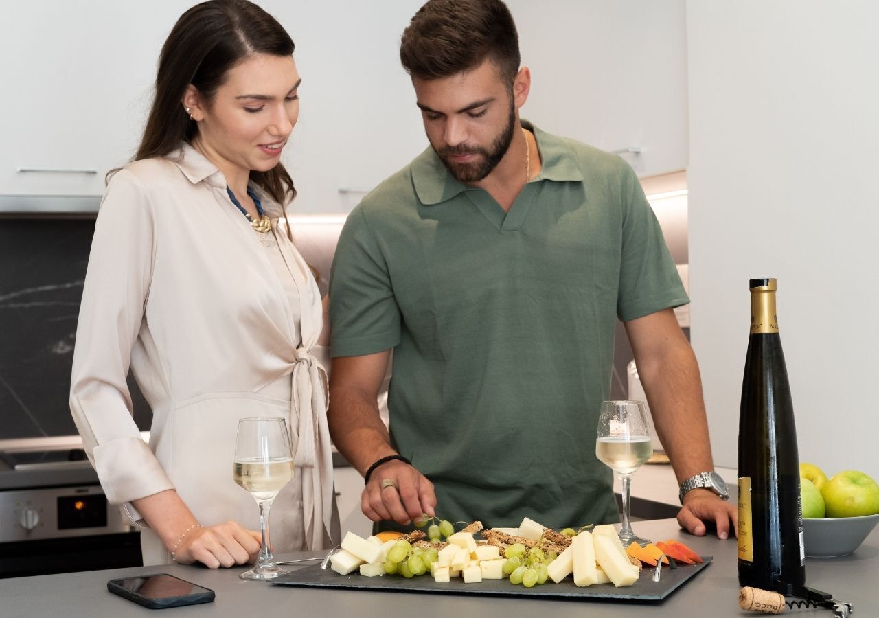 Un uomo e una donna in piedi a un tavolo con un tagliere di formaggi e frutta, calici di vino e una bottiglia.