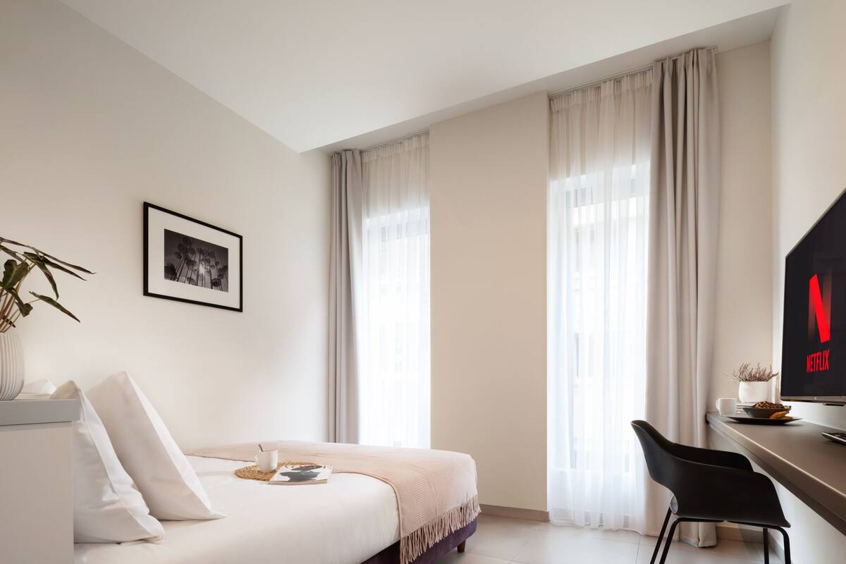 A modern hotel room with a bed, a desk with a chair, a television, and a window with curtains.