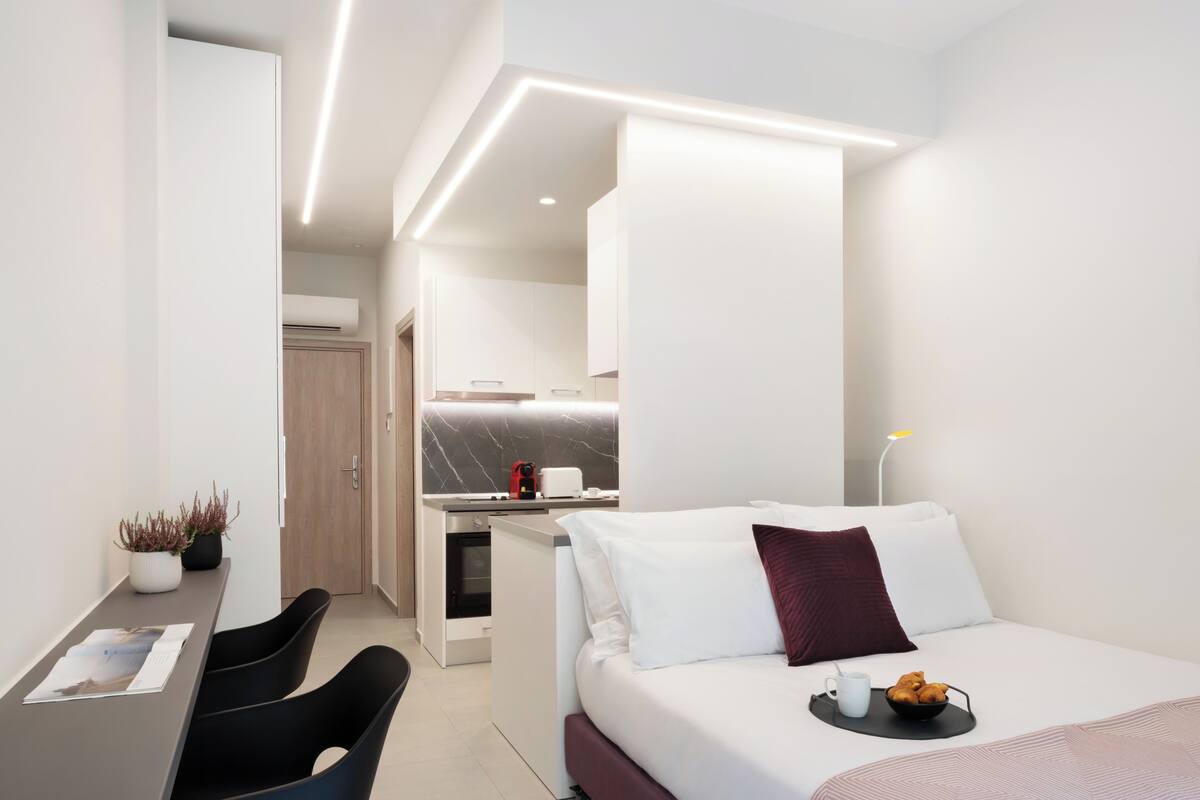 A modern hotel room with a bed, a desk, a chair, a small kitchen area, and a dining table.