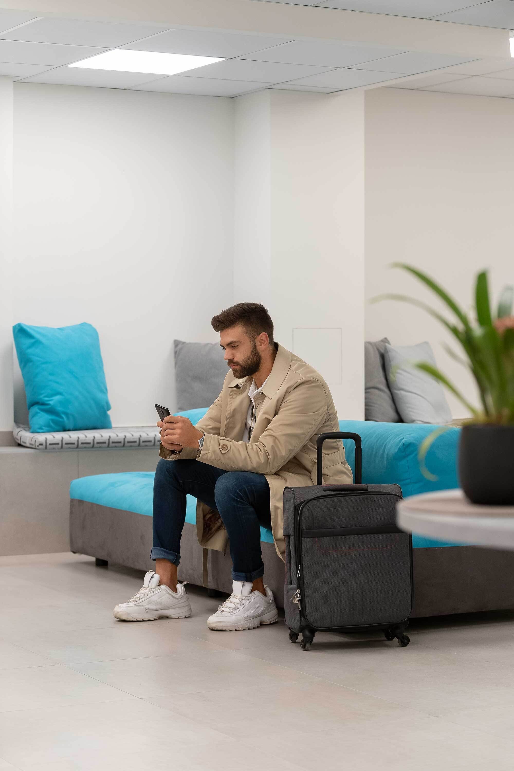 Una persona seduta su un divano grigio con un cuscino blu, che tiene in mano uno smartphone, in una stanza con una valigia grigia e una pianta in vaso.