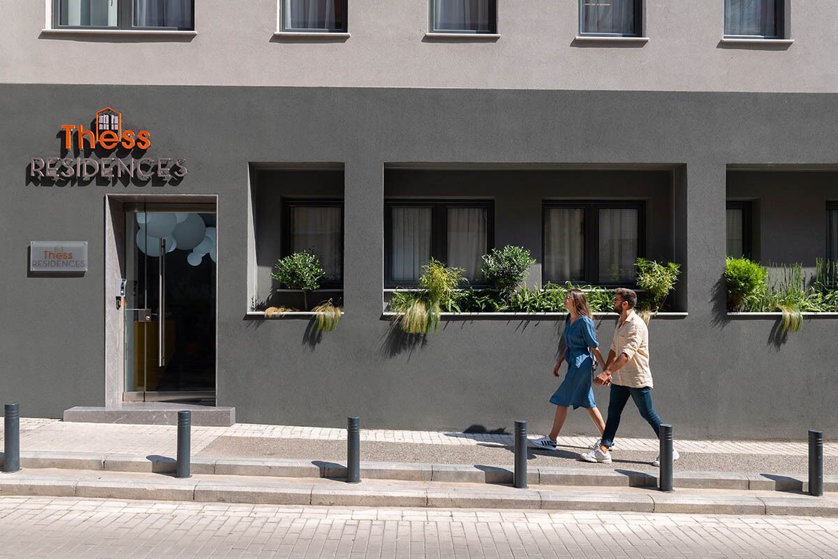 Un edificio moderno con un letrero que dice "Thess Residences". Presenta una fachada limpia y minimalista con ventanas amplias y pequeños maceteros en los alféizares.