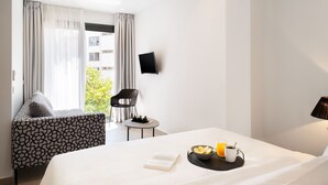 A hotel room with a bed, a small table with a bowl of fruit and a cup, a chair, a TV on the wall, and a window with curtains. - Thess Residences (Thessaloniki)