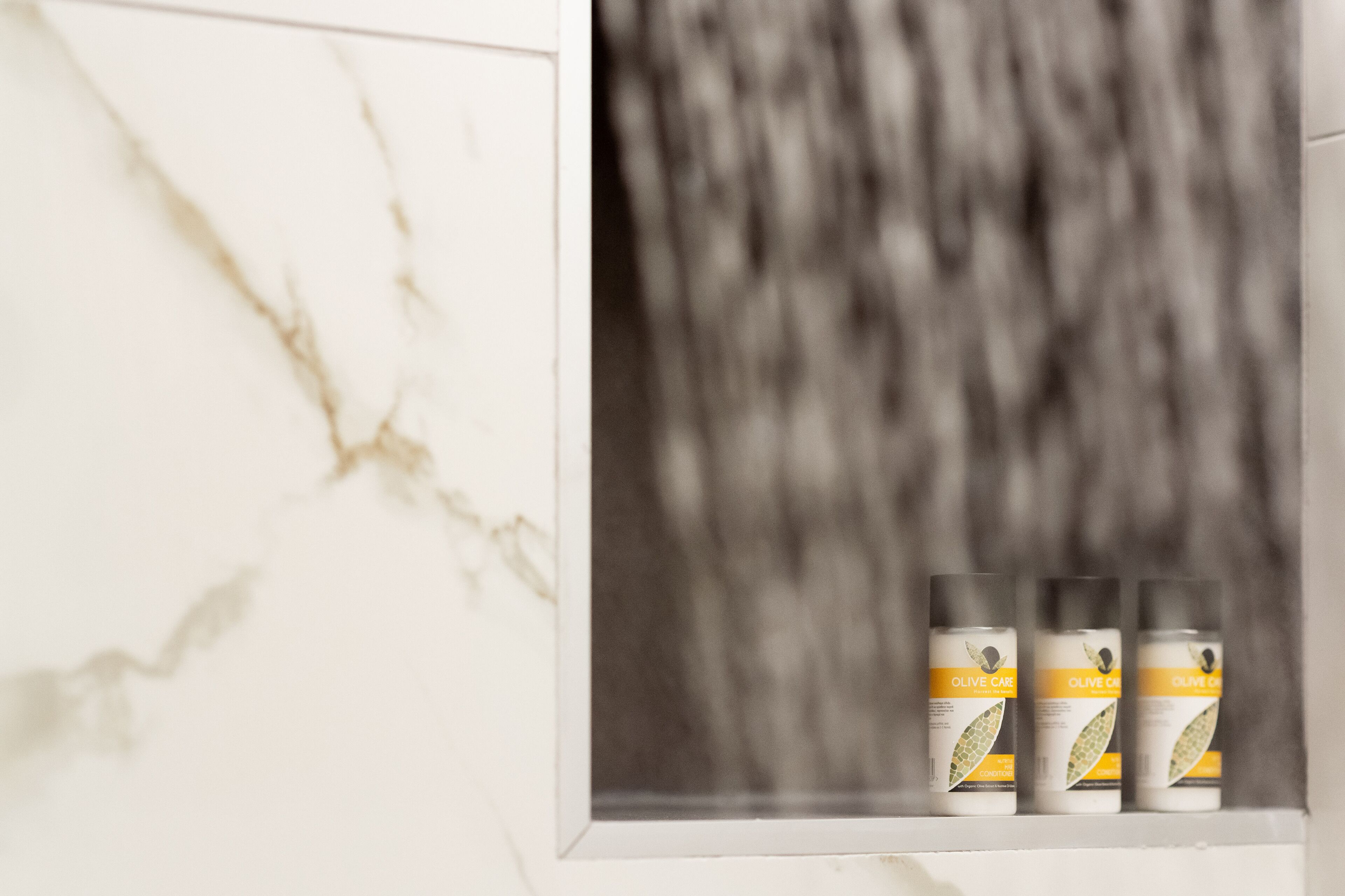 A bathroom with a marble wall and three bottles of olive oil on a shelf.