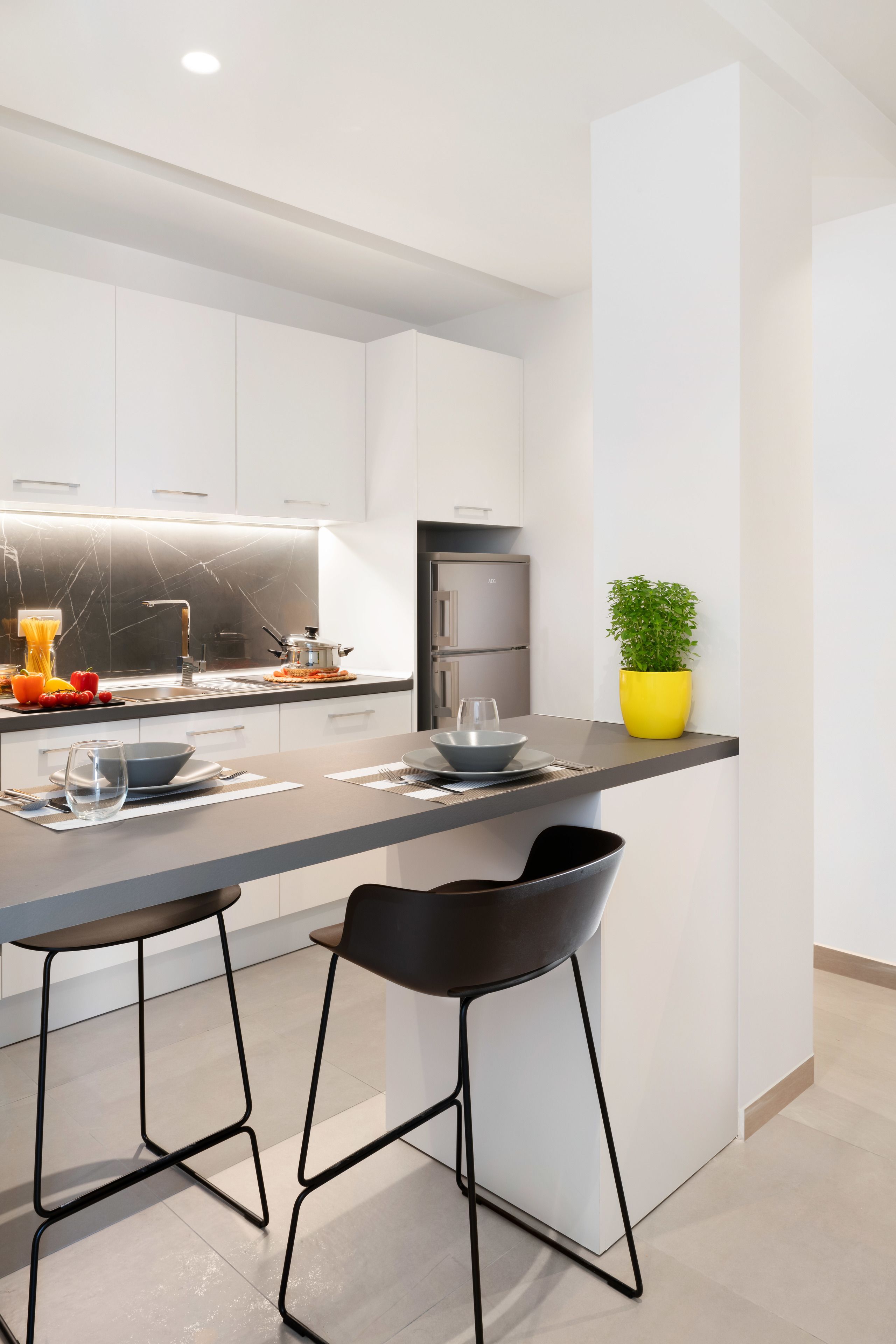 A modern kitchen with white cabinets, a grey countertop, two black bar stools, a yellow plant, and a stainless steel refrigerator.