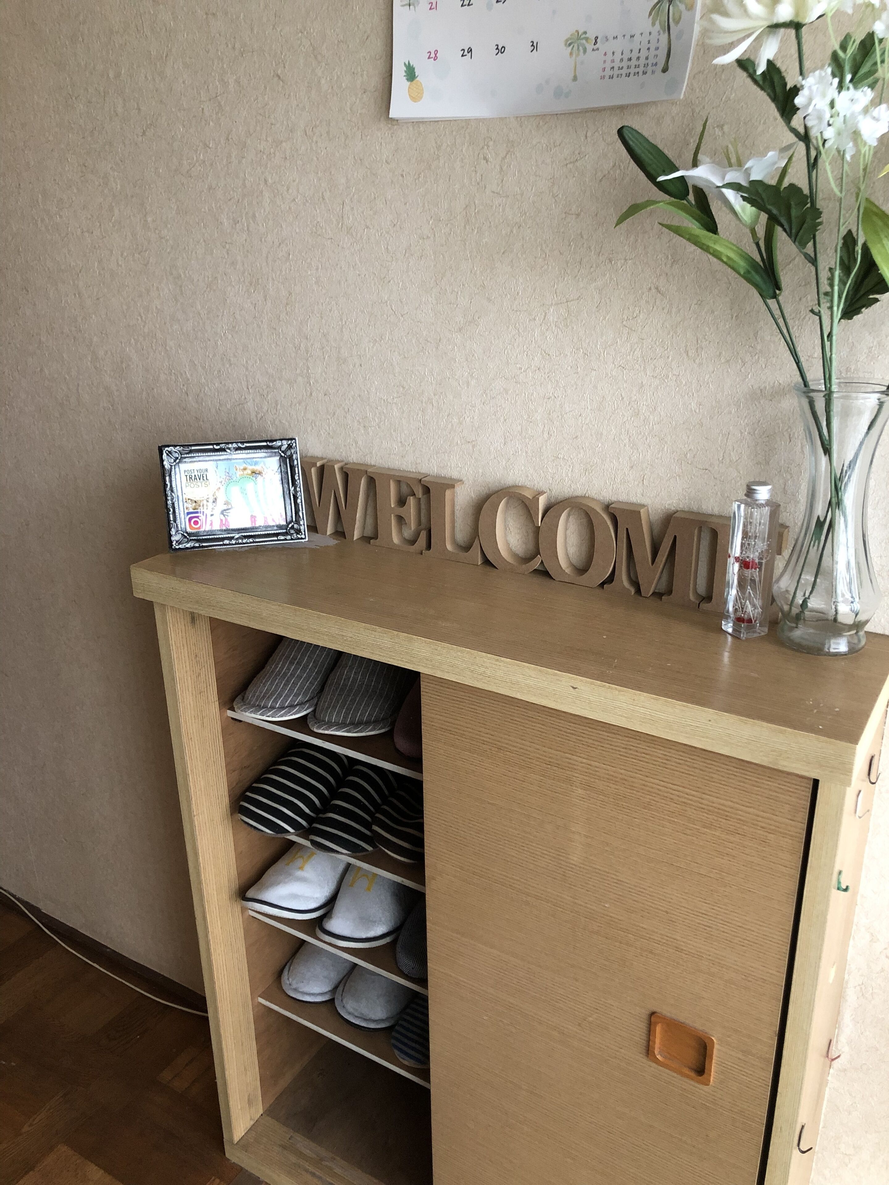 A wooden welcome sign with a calendar and a vase with flowers on a shelf.