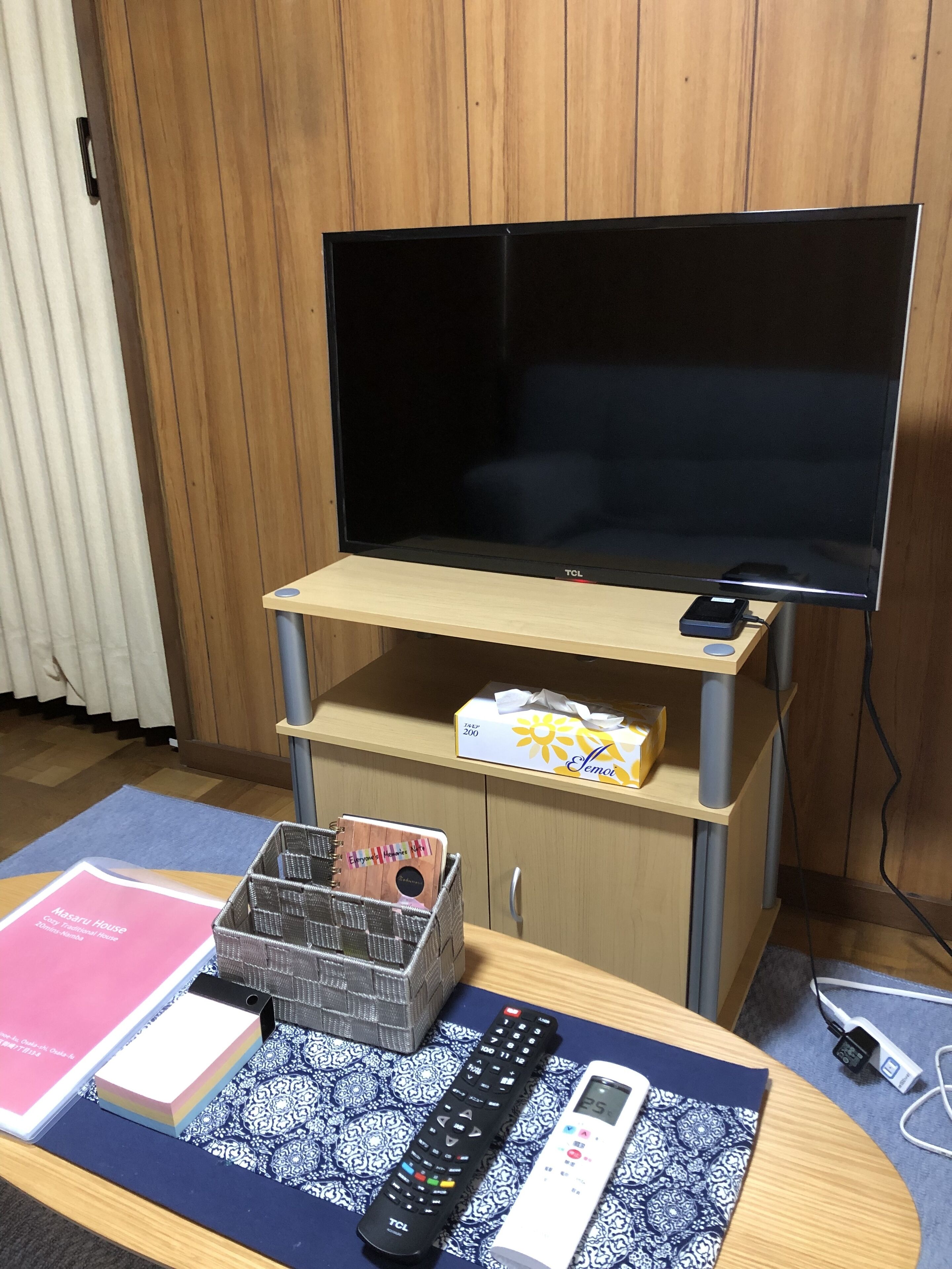 Ein Zimmer mit Fernseher auf einem Holzboden, einer Fernbedienung, einer digitalen Uhr und einer Tasche Taschentücher.