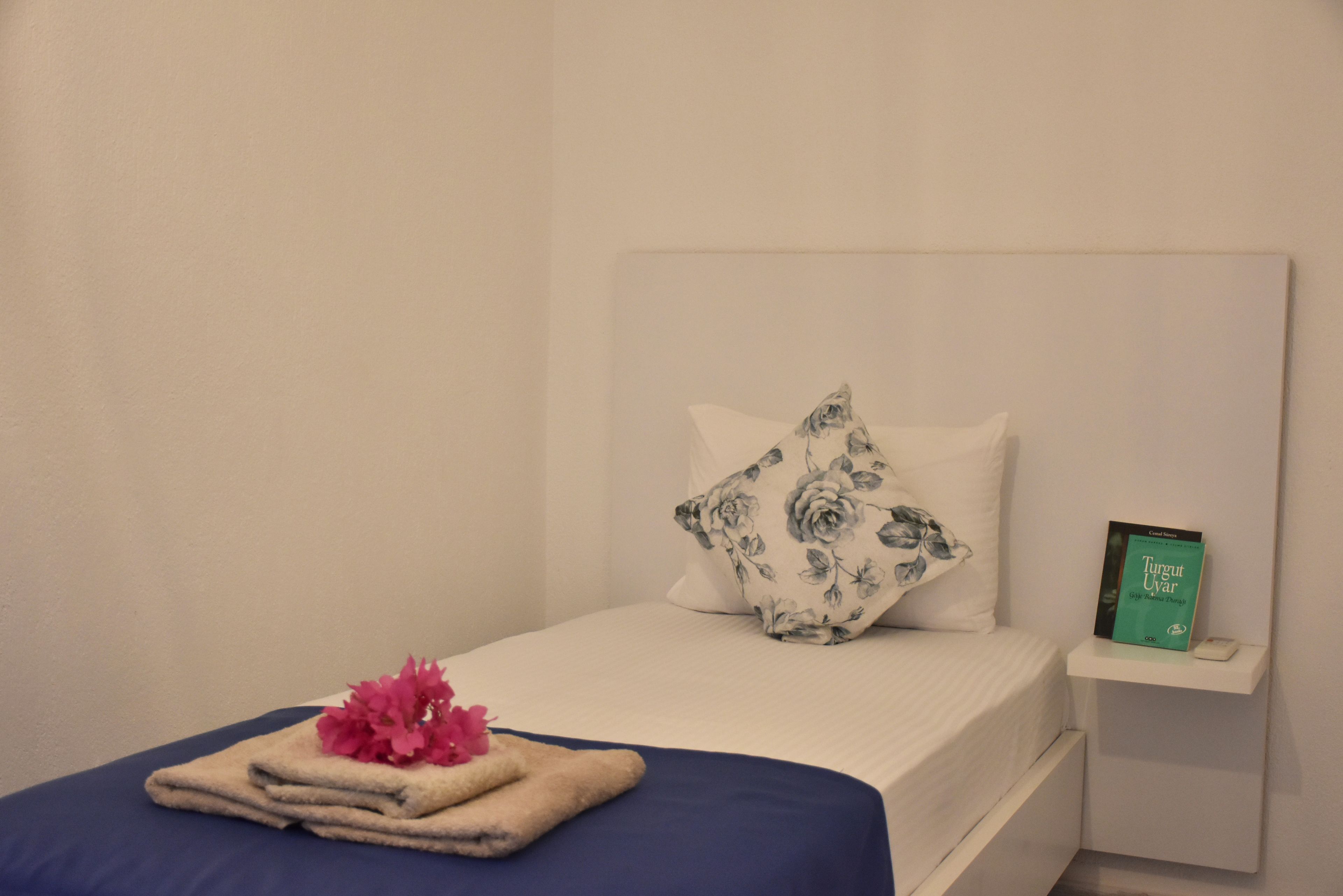 A neatly made bed with a blue bedspread and a floral pillow, a small bedside table with a book, and a pink flower arrangement on the bed.