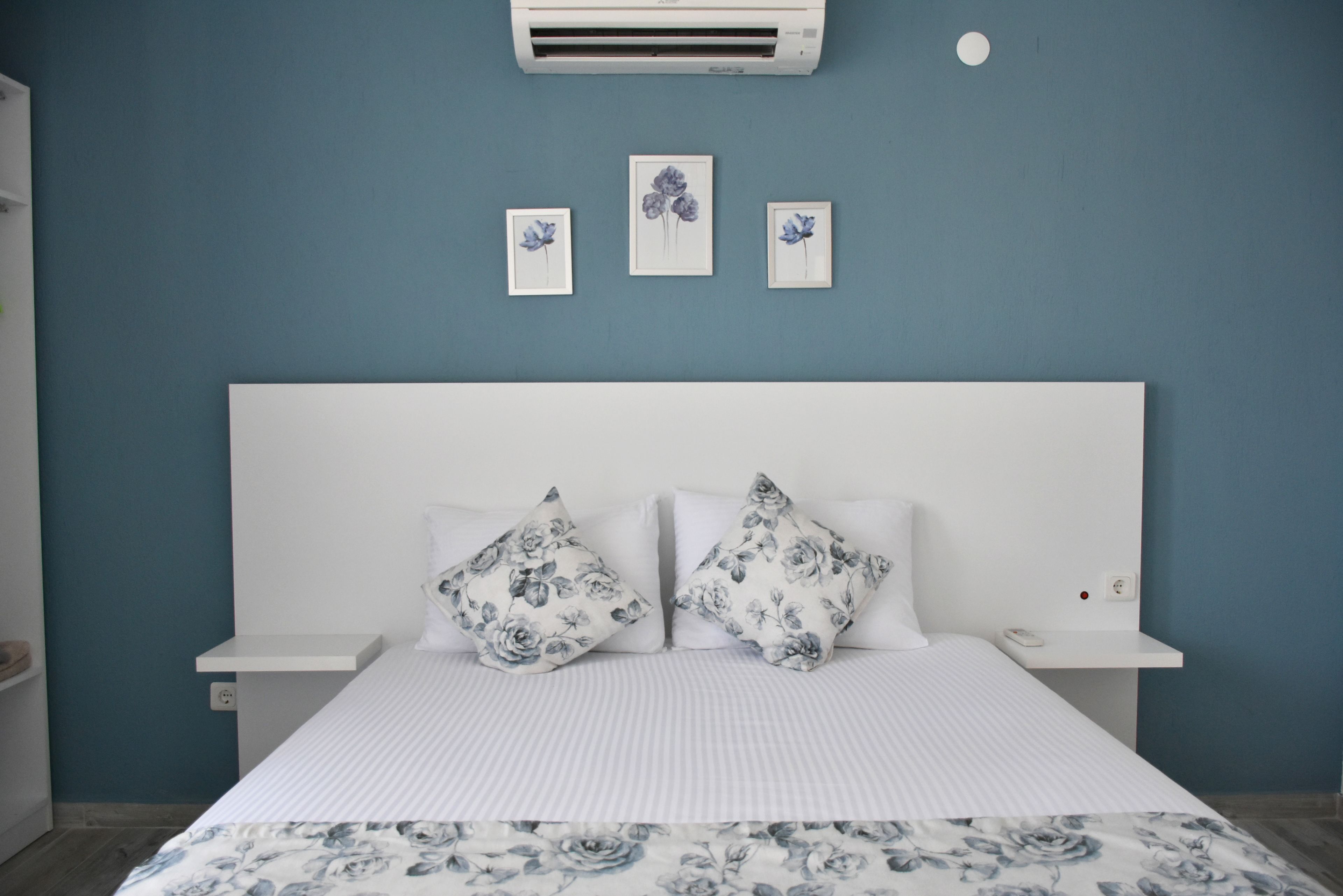A bedroom with a white bed, floral-patterned pillows, a blue wall, and a white nightstand with a phone.