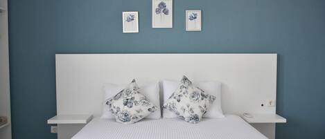 A bedroom with a white bed, floral-patterned pillows, a blue wall, and a white nightstand with a phone.