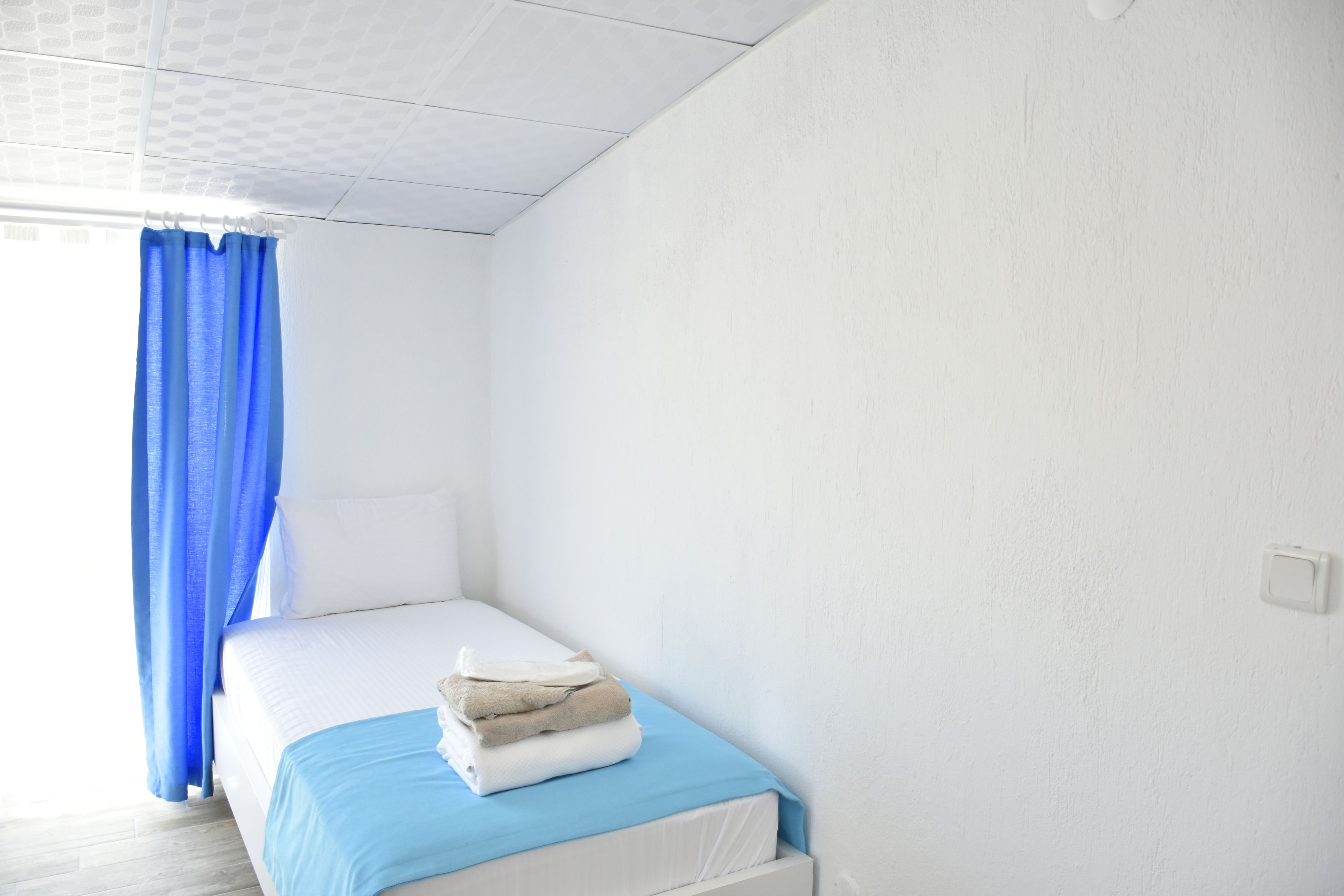 A small, single-bed room with a white bed, a blue bedspread, and a stack of folded towels on the bed.
