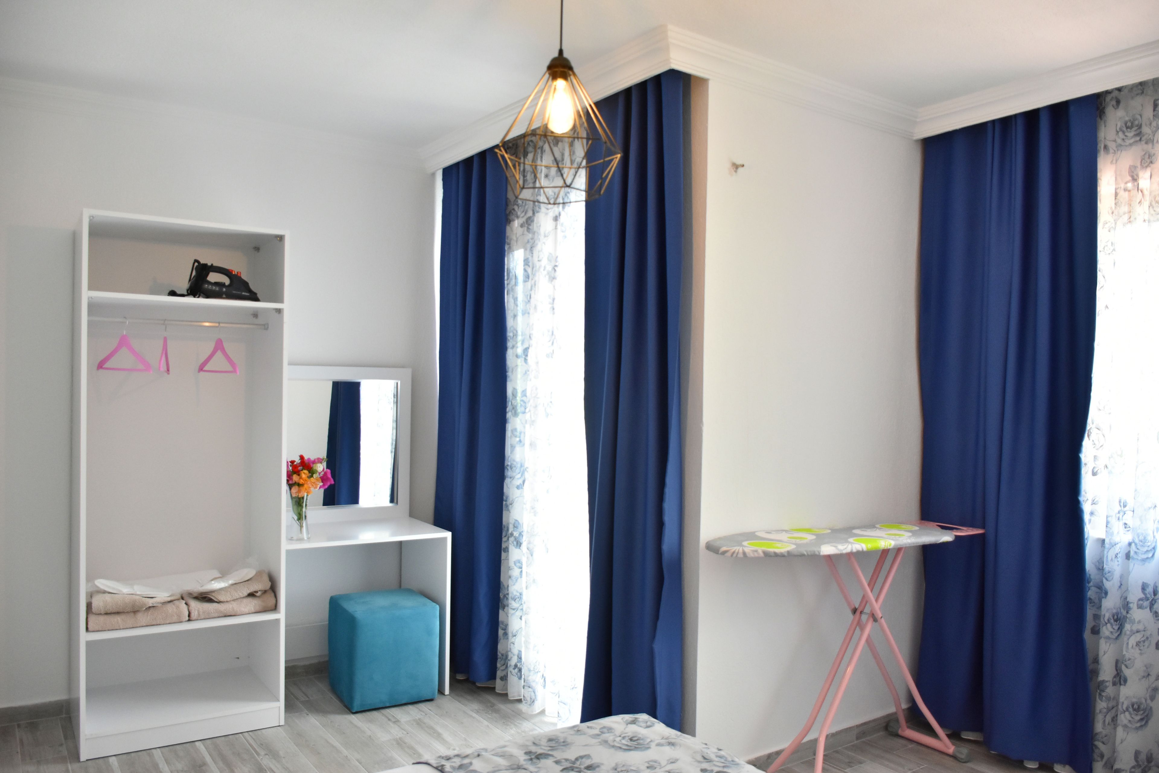 A bedroom with a wardrobe, a vanity with a mirror, a blue ottoman, a pink hanger rack, and a small ironing table.