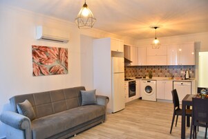 Cocina compacta con muebles blancos, horno empotrado y lavadora. En la sala de estar, un sofá gris con cojines.