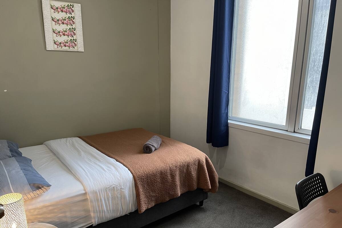 Un dormitorio pequeño y ordenado con cama, lámpara de noche, una silla y ventana con cortinas.