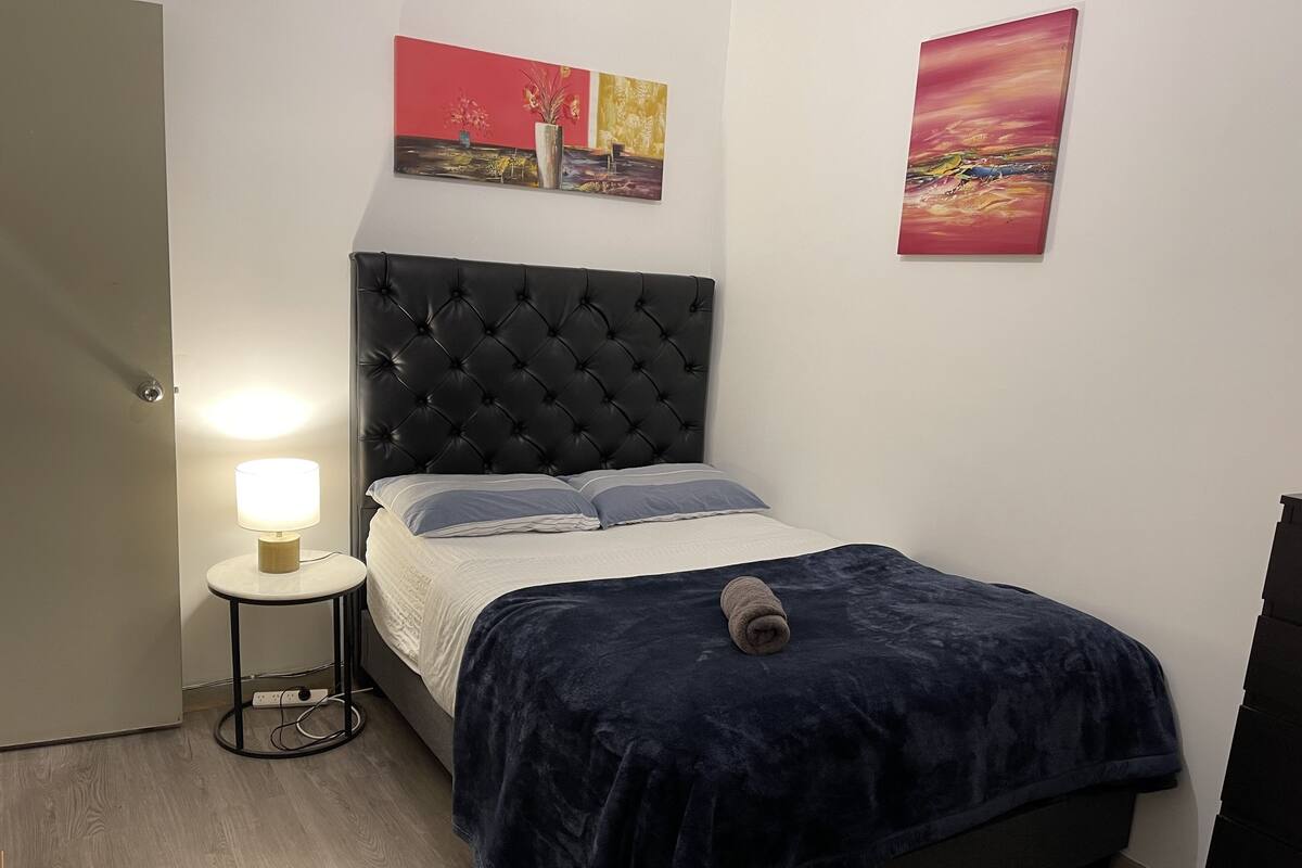 Un dormitorio con cabecera capitonada, una mesita de noche con lámpara, una cama con manta azul oscuro y dos cuadros en la pared.