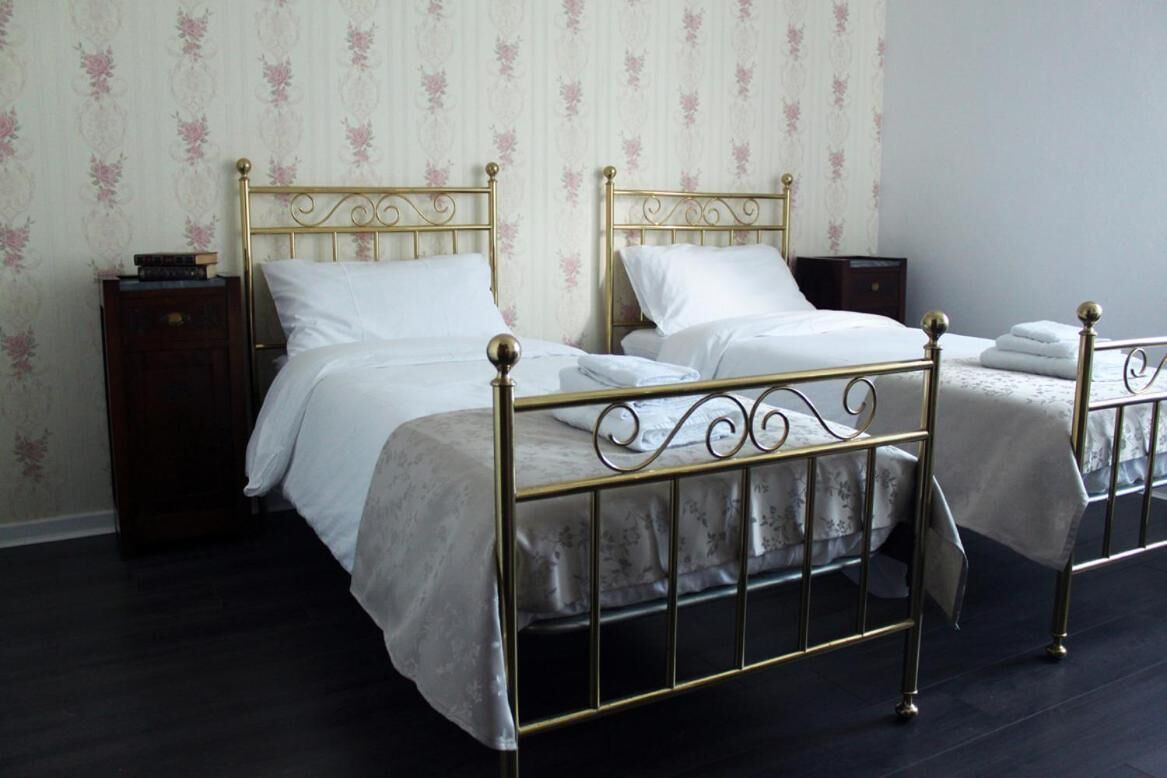 Une chambre avec deux lits simples, chacun doté d’un cadre en métal et de draps blancs. Une petite table de chevet en bois, sur laquelle se trouvent un livre et une lampe, est placée à côté de l’un des lits. Les murs sont recouverts d’un papier peint à motifs floraux.