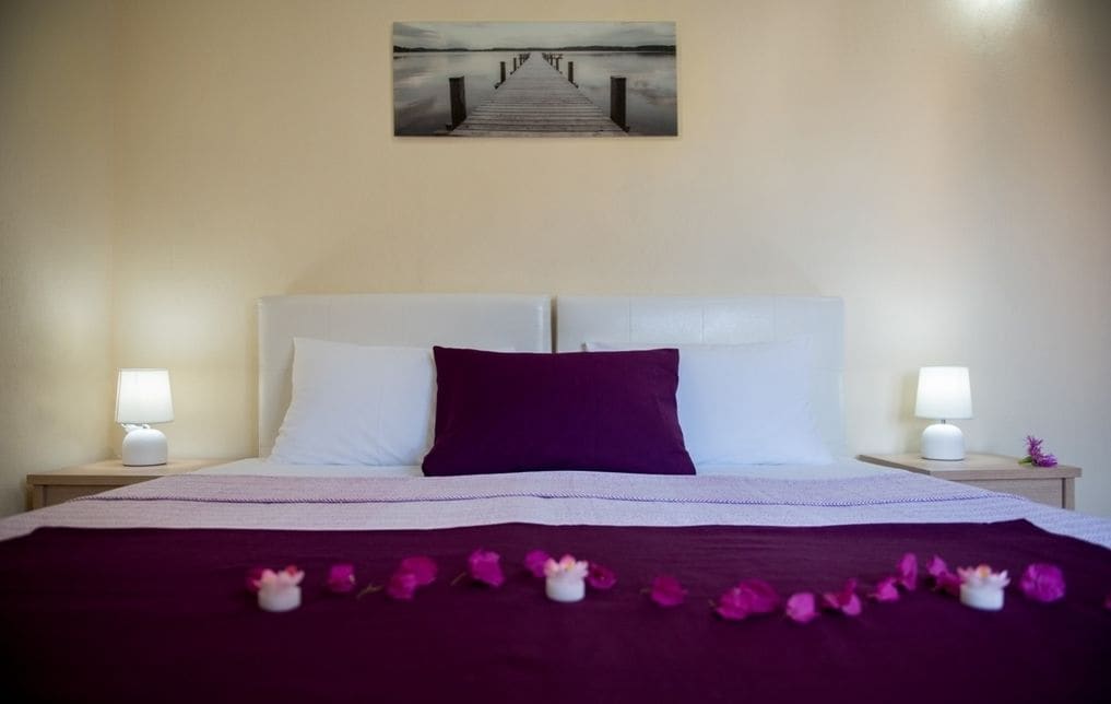 Un lit bien fait, avec un oreiller violet, des oreillers blancs et une couvre-lit violette. Des pétales de fleurs sont disposés sur le lit.
