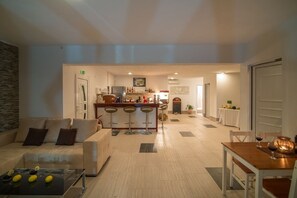 A modern living room with a sofa, a coffee table, and a dining area with a table and chairs. There is a kitchen area with a bar and stools in the background. - Caretta Village (Sithonia)