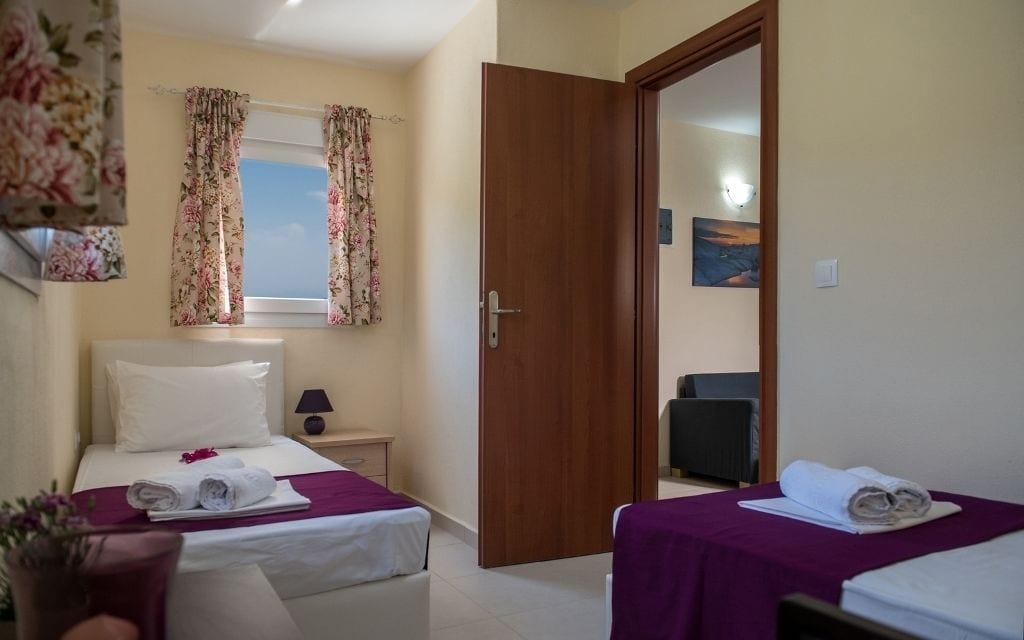 Une chambre d’hôtel avec deux lits, une fenêtre ornée de rideaux à motifs floraux, une porte en bois, une petite table de chevet avec une lampe et un tableau accroché au mur.