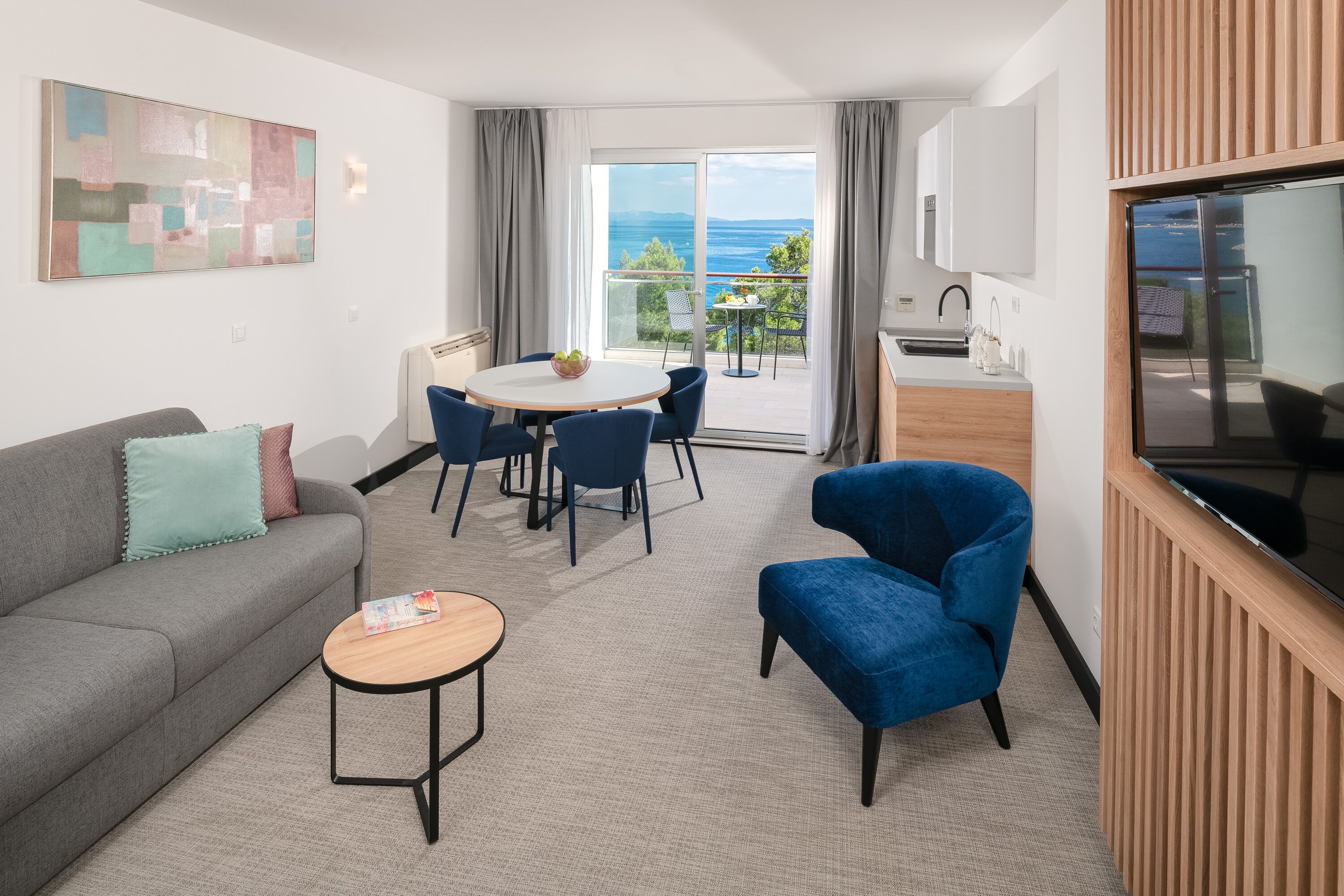 En moderne stue med en grå sofa, lænestol i blåt og et rundt bord med frugtskål. En balkon med havudsigt ses gennem glasdørene.