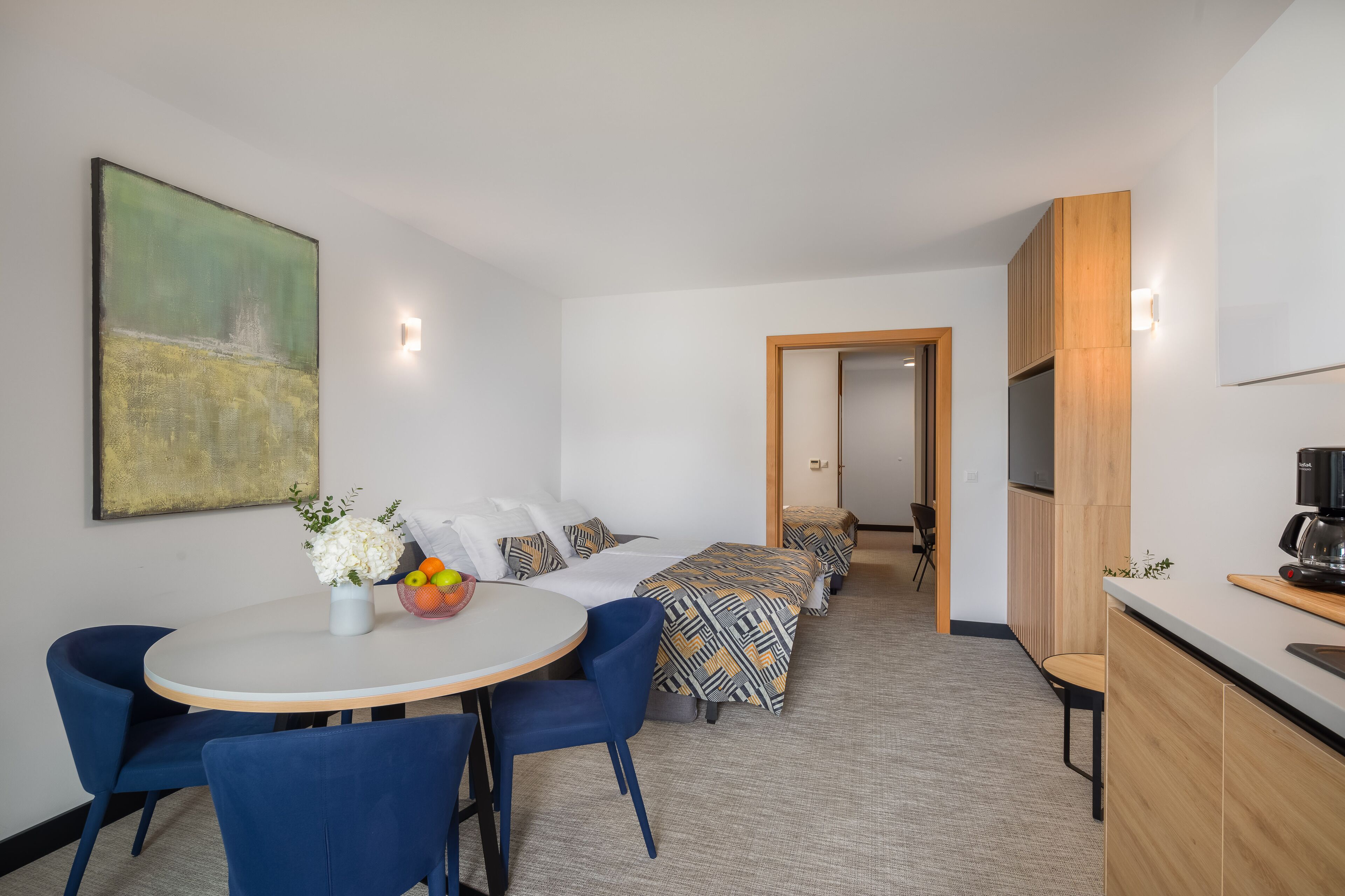 A modern hotel room with a dining area, a bed with patterned bedding, a large abstract painting, and a kitchenette.