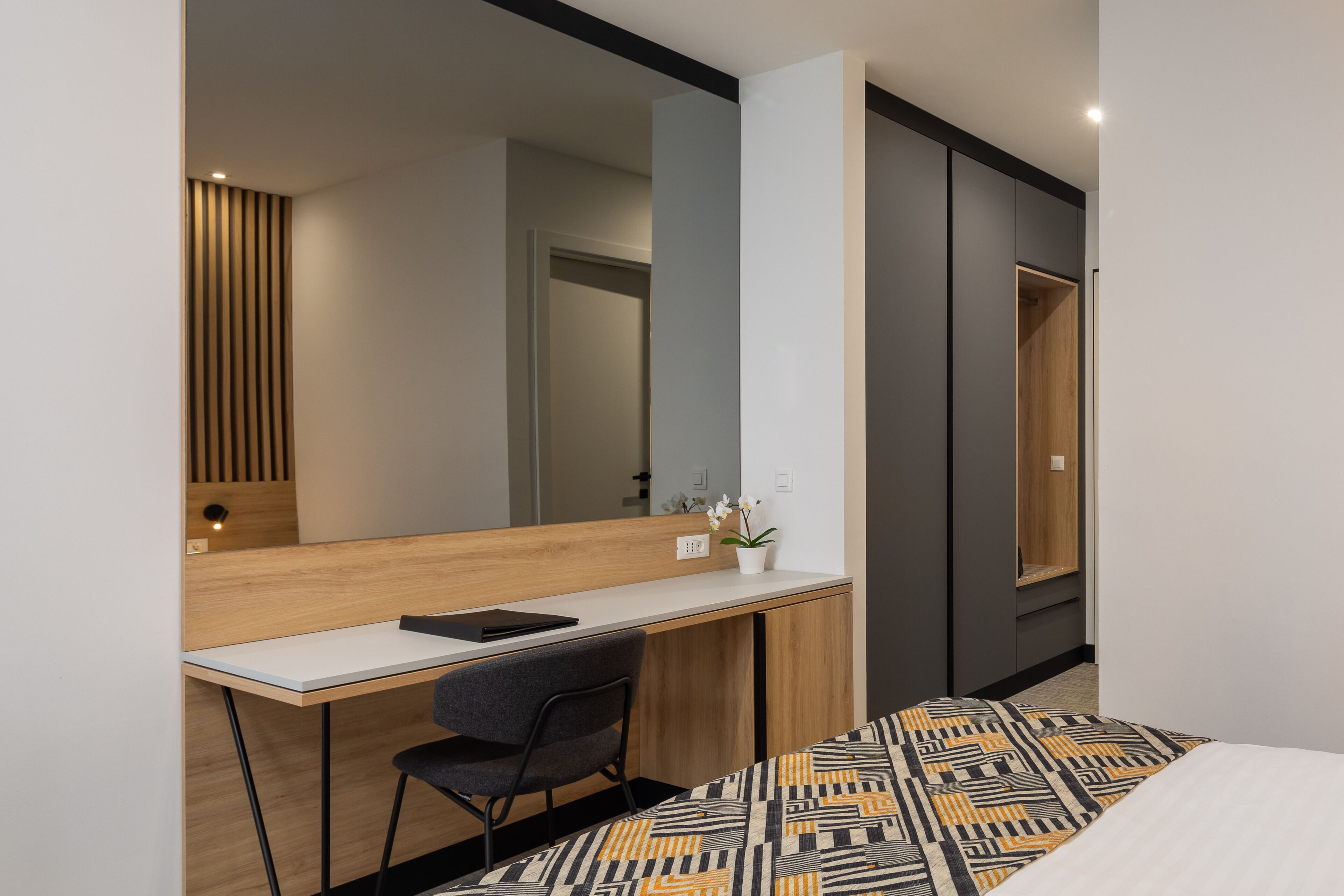 Una habitación de hotel moderna con un escritorio de madera, una silla, una cama con ropa de cama estampada, un espejo grande y un armario.