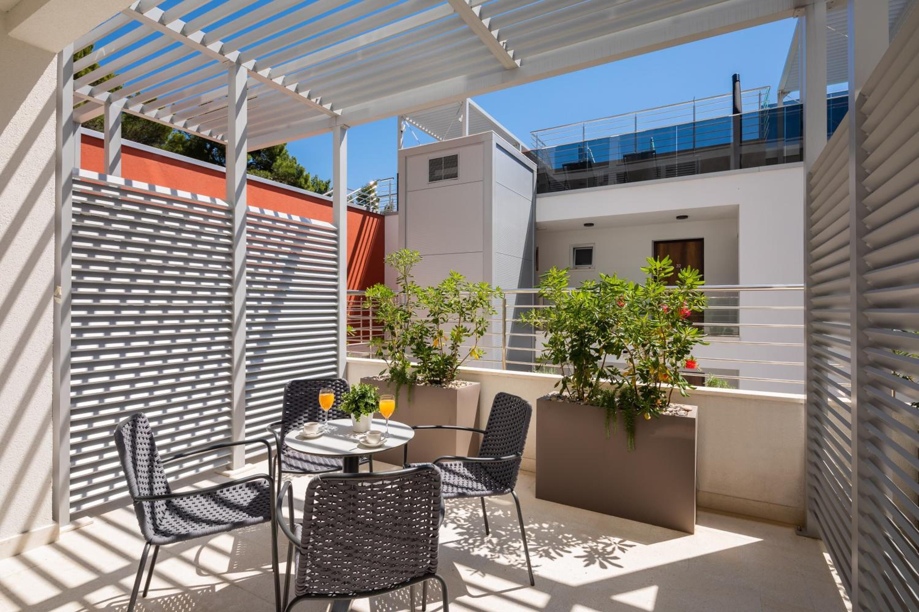 Un patio exterior con una mesa redonda y sillas, rodeado de plantas en macetas y una pérgola blanca.