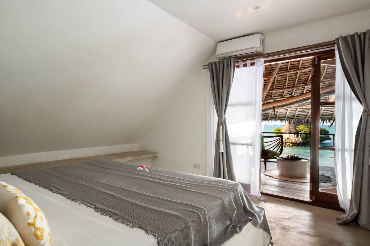 Ein Schlafzimmer mit einem Bett, Blick auf einen Balkon mit einem Strohdach und einem Blumentopf.