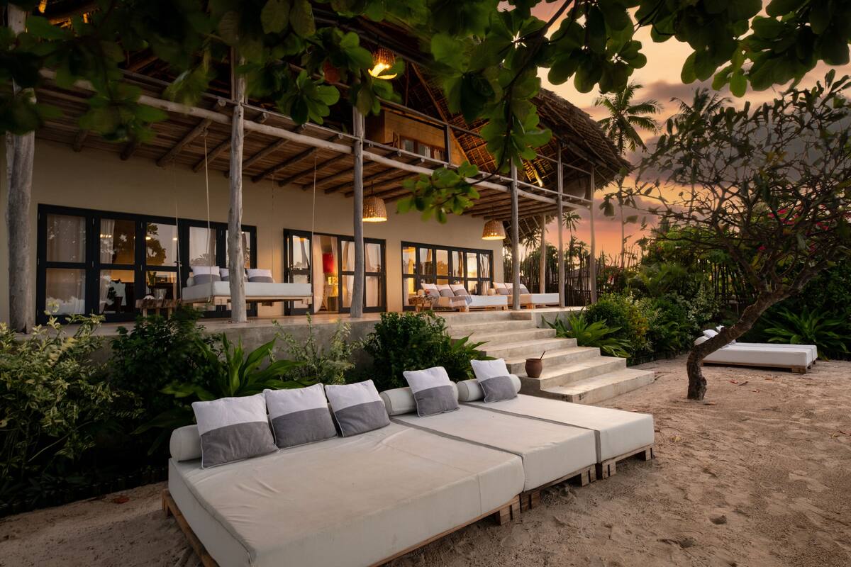 Ein Strandresort mit einer Lounge, die ein langes Sofa mit Kissen, eine Treppe zu einer überdachten Terrasse und üppiges Grün bietet.