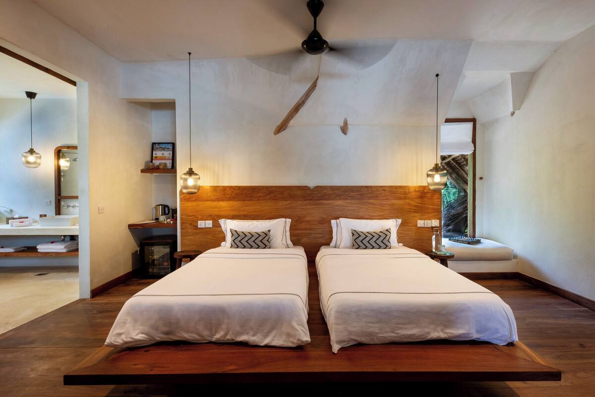 Ein modernes Hotelzimmer mit zwei Betten, einem Holzkopfende, Pendelleuchten und einem Deckenventilator.