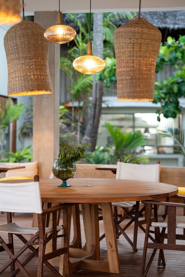 Una mesa de comedor de madera con lámparas colgantes de mimbre, rodeada de sillas, en una habitación con vistas a la vegetación.