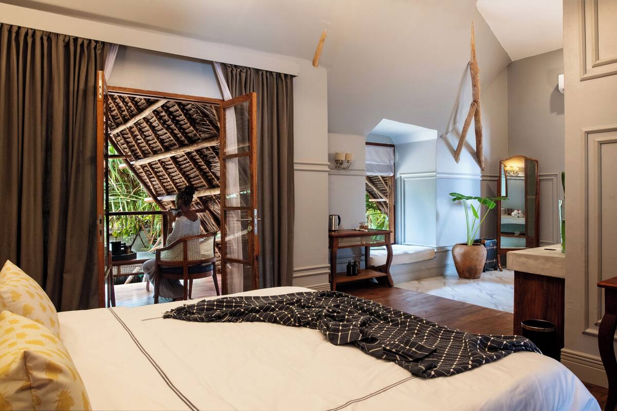 Ein Hotelzimmer mit einem großen Bett, einem Holztisch, einem Stuhl, einer Topfpflanze und Blick ins Freie.