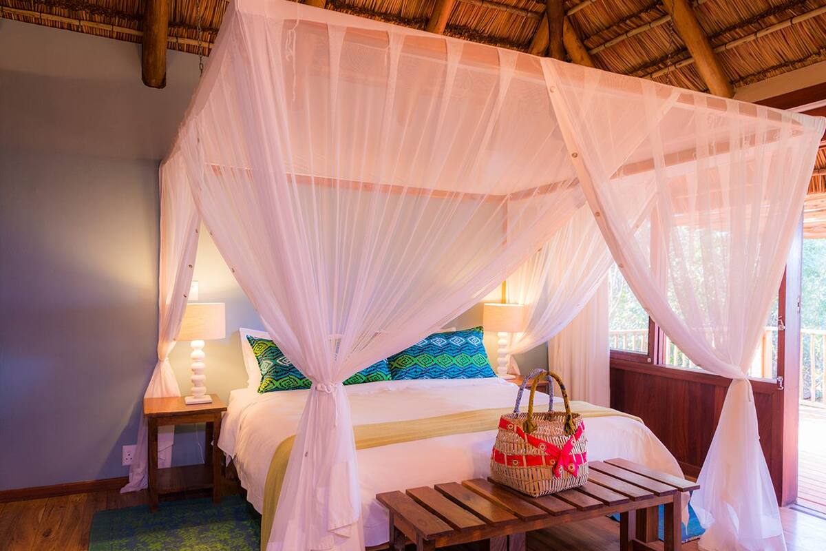 A room with a bed under a canopy, a wooden nightstand, a lamp, and a wicker basket on a wooden table.
