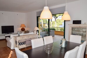 Un salon moderne comprenant un téléviseur, un canapé, une table à manger et des chaises. Il y a un balcon avec vue sur la verdure.