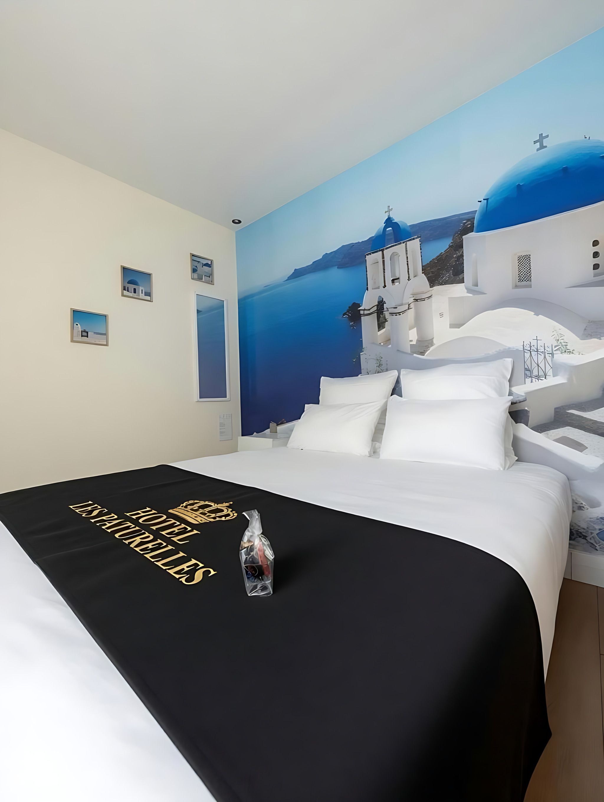 Une chambre d’hôtel avec un lit, une couvre-lit noire ornée de motifs dorés, une bouteille et une fresque représentant un bâtiment blanc surmonté d’un dôme bleu.