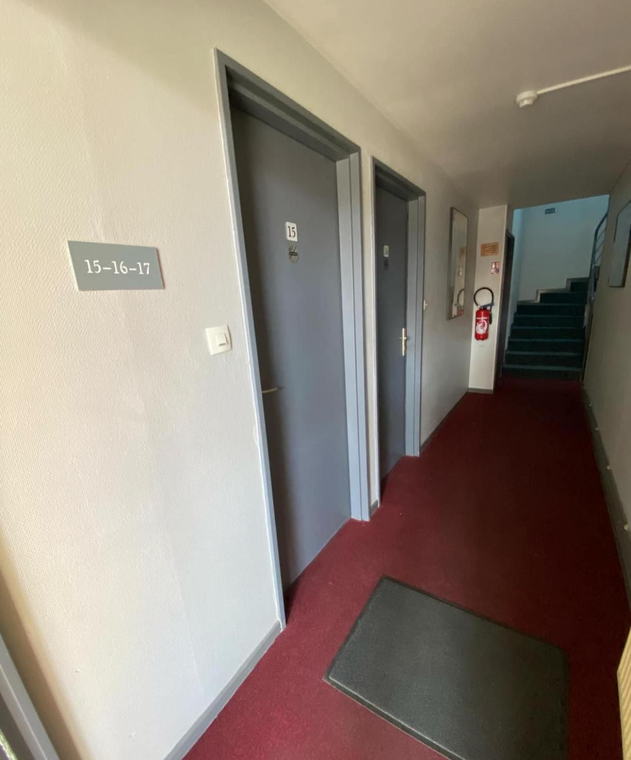 Un couloir avec des portes numérotées 15, 16 et 17, un extincteur et une moquette rouge.