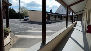 A covered outdoor area with a tiled floor, a white building in the background, and a parked car. - Hôtel Les Paturelles  (Avesnelles)