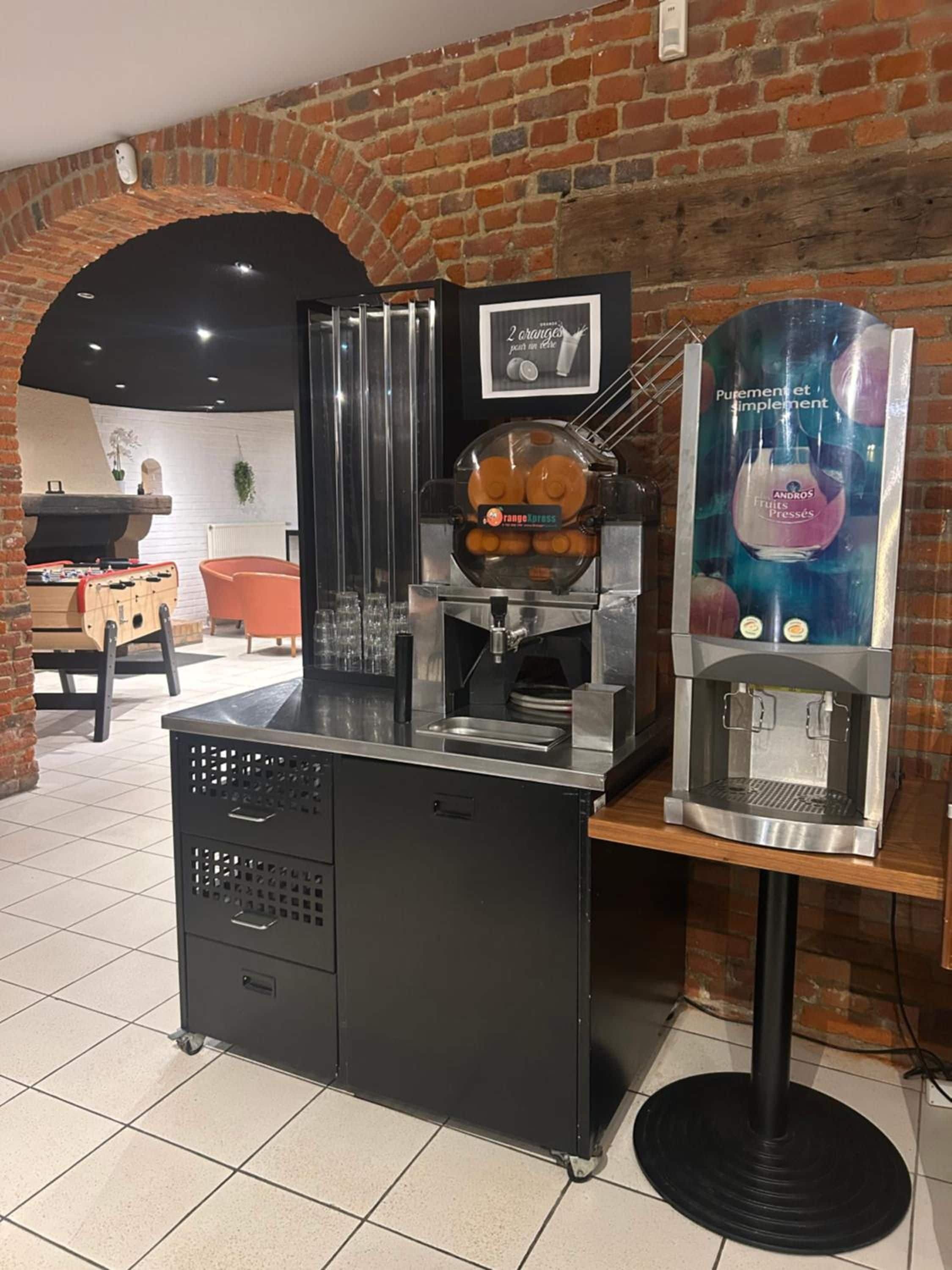 A coffee machine and a juice dispenser in a cafe setting with a brick wall and a foosball table in the background.
