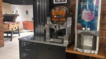 A coffee machine and a juice dispenser in a cafe setting with a brick wall and a foosball table in the background.