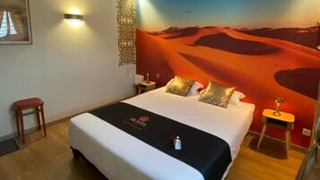Habitación de hotel con una cama grande, una mesita de noche, una pequeña mesa, una silla, una lámpara de pared y un mural al estilo del desierto en la pared.