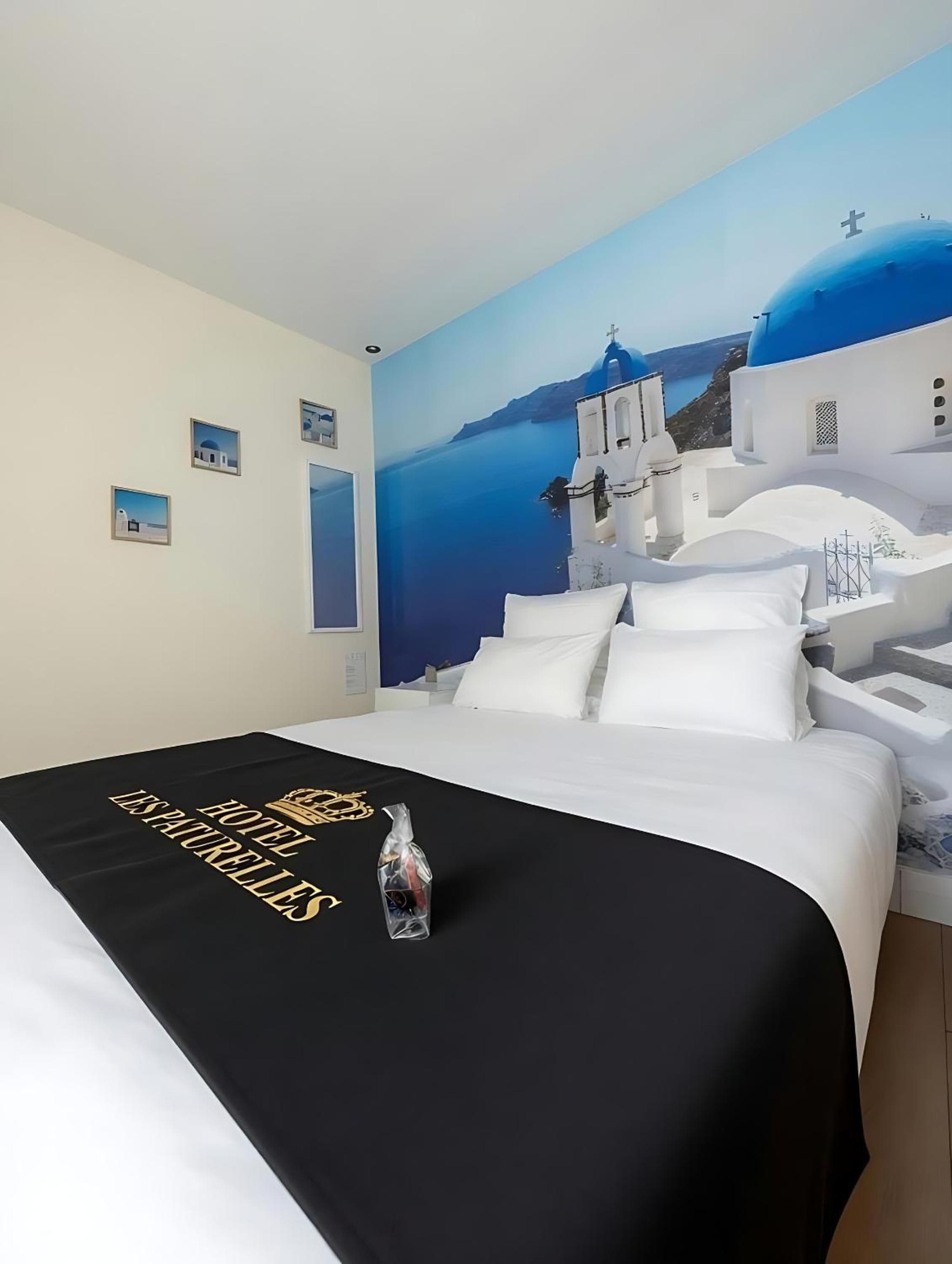 Una habitación de hotel con cama, una colcha negra con letras doradas, una botella y un mural en la pared de un edificio blanco con cúpula azul.