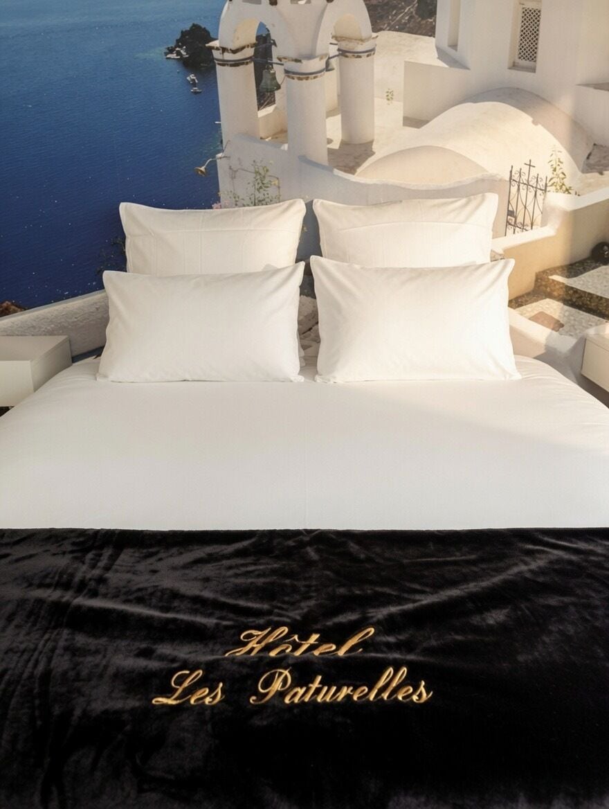 Cama con ropa de cama blanca y un plaid de terciopelo negro, con vistas a la costa, edificios blancos y mar azul.