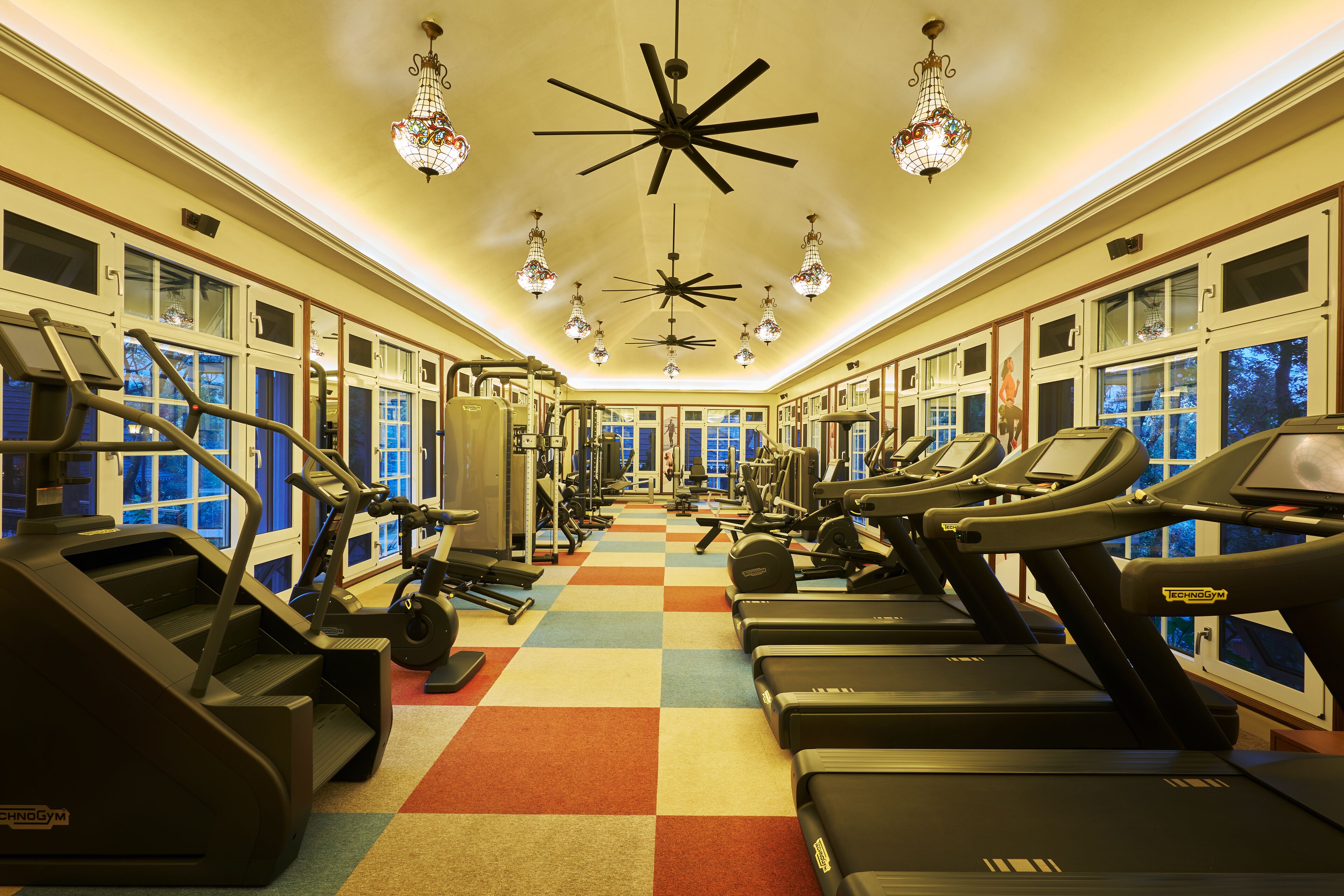 Un gimnasio con cintas de correr, equipos de ejercicio y ventanas amplias.