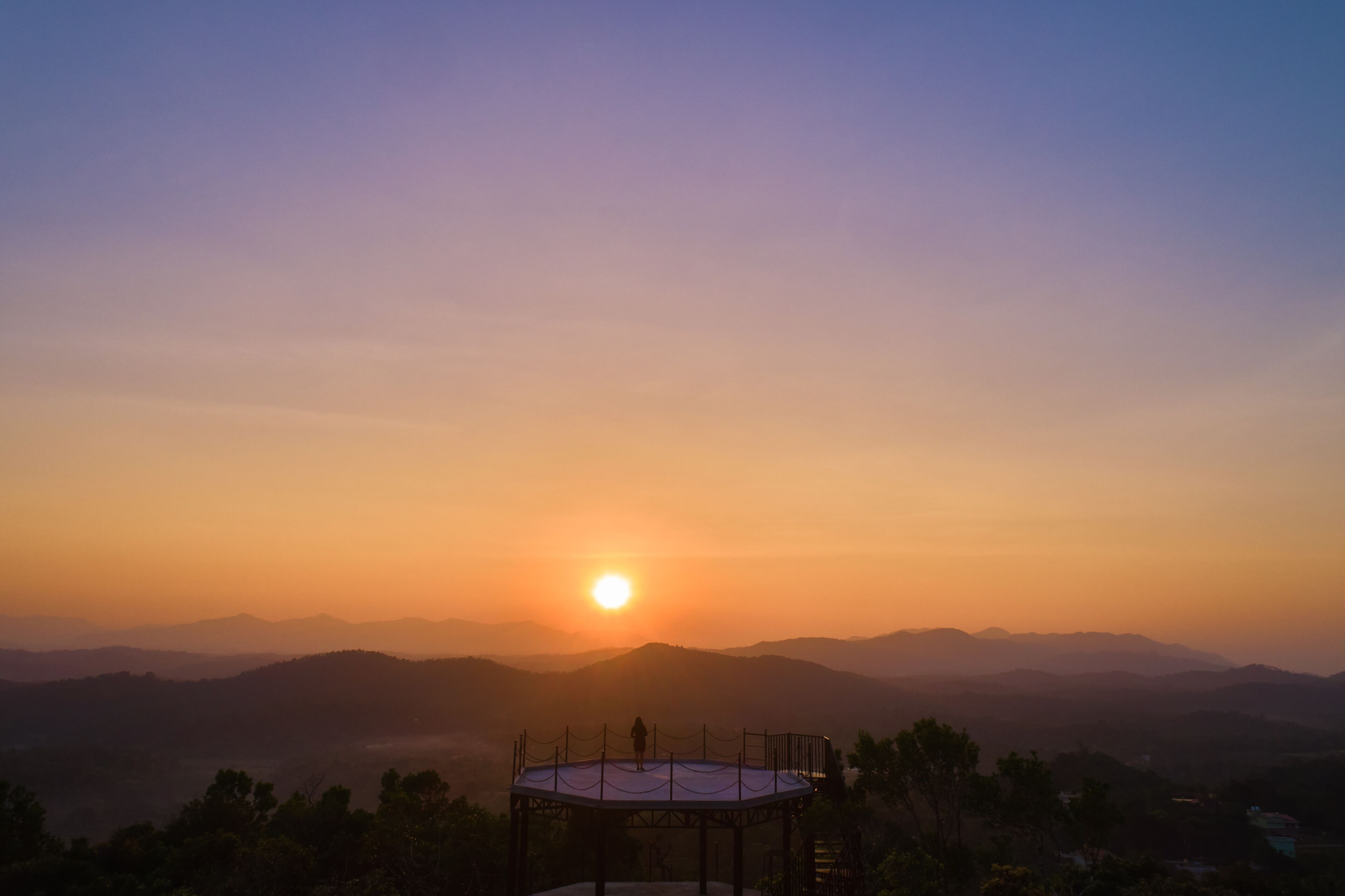 Vista del atardecer desde una plataforma de observación circular con una persona de pie, contemplando una cadena montañosa.